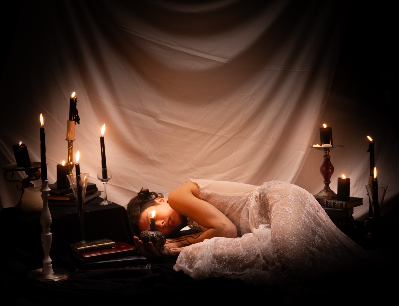 Tori Louise asleep on the floor in a white lace dress, her head beside a lit candle, among candlesticks and stacked books against a draped sheet.